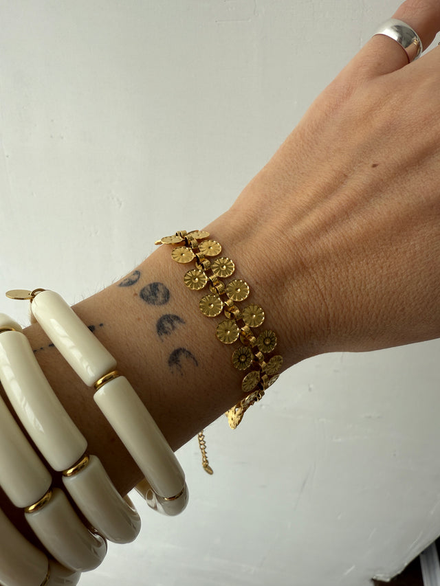 Pulseira dourada em aço inoxidável, elegante e durável, adornando um pulso com tatuagens e pulseiras brancas.