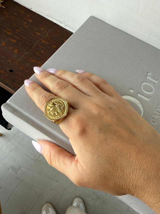 Close-up of a hand wearing a thick gold ring with a coin design, beside a stylish Dior box.