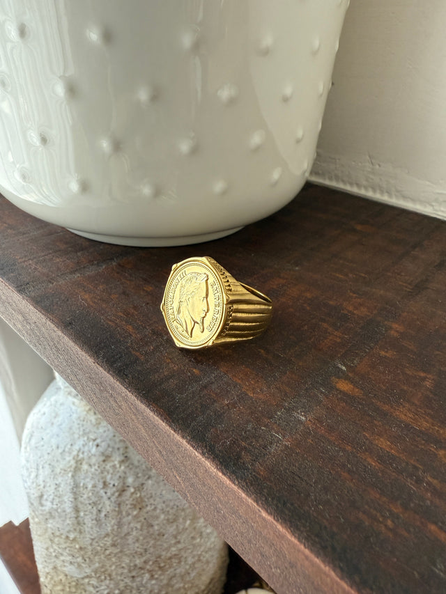 Anel Grosso Com Moeda em ouro sobre uma prateleira de madeira, destacando o design elegante e moderno.
