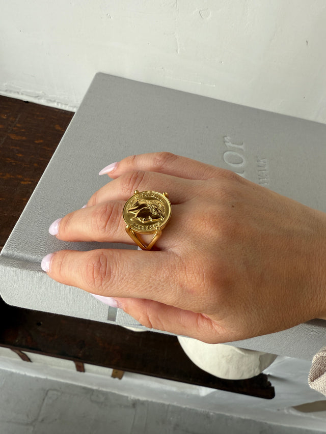 Elegante anel com moeda dourada, exibido na mão sobre uma superfície clara, destacando seu design robusto.