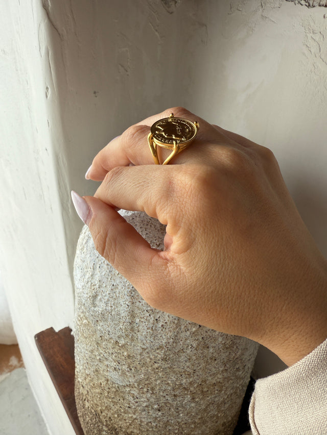 Elegante anel robusto em ouro com moeda, exibido em mão feminina sobre superfície texturizada.