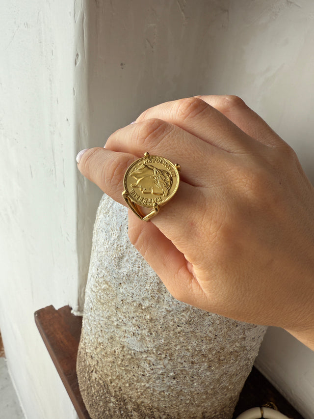 Anel robusto e elegante com moeda dourada, ajustável, exibido em mão feminina.