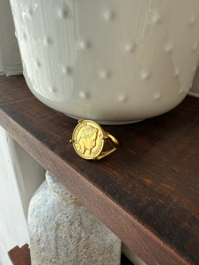 Anel robusto e elegante com moeda, exibido sobre uma prateleira de madeira, ideal para qualquer estilo.