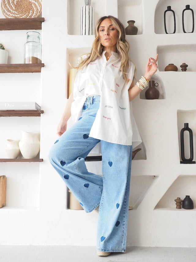 Woman wearing oversized shirt with embroidered words and heart-patterned jeans, posing in a stylish interior.