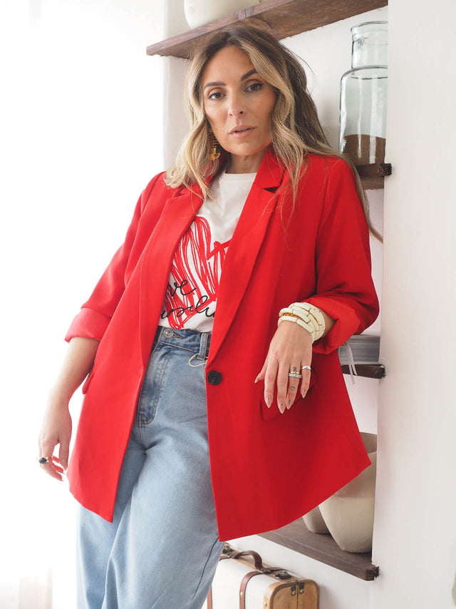 Modelo vestindo blazer vermelho de corte reto, combinado com camiseta estilizada e jeans, em ambiente iluminado.