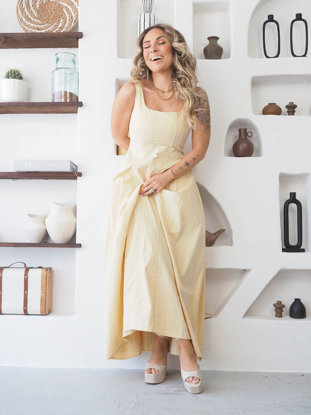Modelo sorrindo usando vestido comprido de alças amarelo em ambiente moderno e elegante.