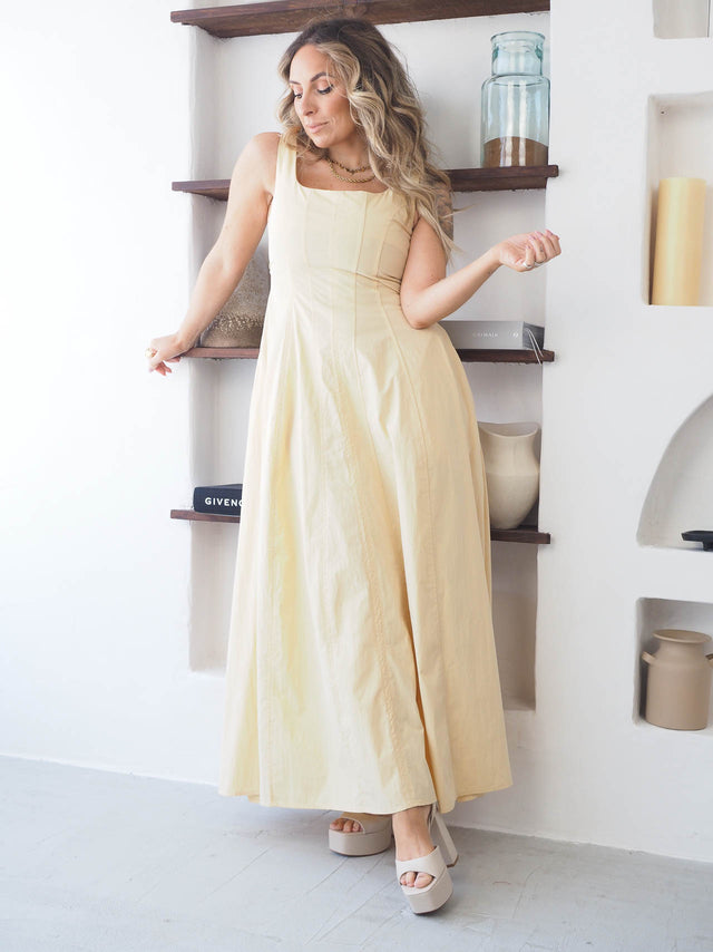 Modelo usando vestido comprido de alças amarelo em ambiente minimalista, destacando a elegância e o corte cintado.