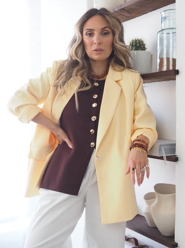 Modelo com blazer básico corte reto amarelo sobre blusa marrom e calças brancas, destacando estilo e elegância.