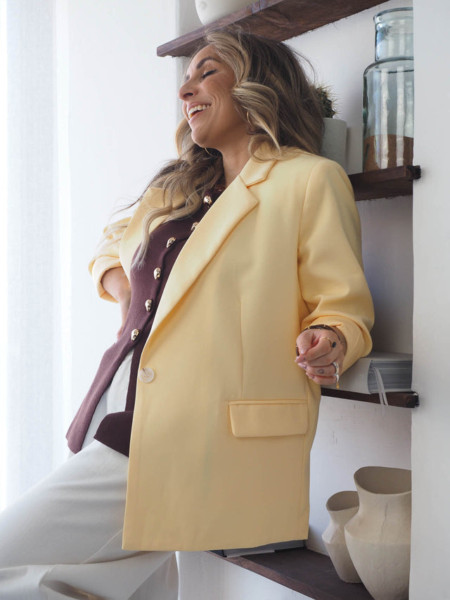 mulher sorridente usando blazer amarelo básico de corte reto em ambiente moderno com prateleiras