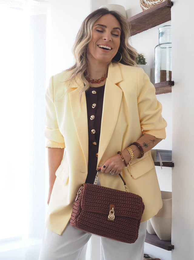 Mulher sorridente usando blazer básico amarelo e segurando bolsa marrom, em ambiente moderno e sofisticado.