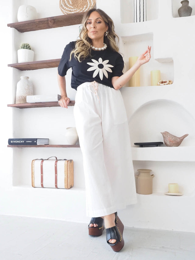 Mulher com t-shirt flor bordada, calças brancas e sandálias, estilo elegante e casual.
