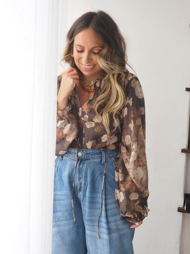 Mulher a usar uma blusa estampada semi transparente com flores, combinada com calças de ganga largas.