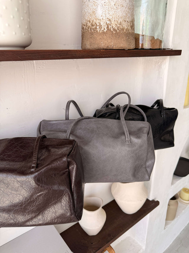 Tote Bag Ecopele em tons de cinza, preto e castanho expostas numa prateleira.