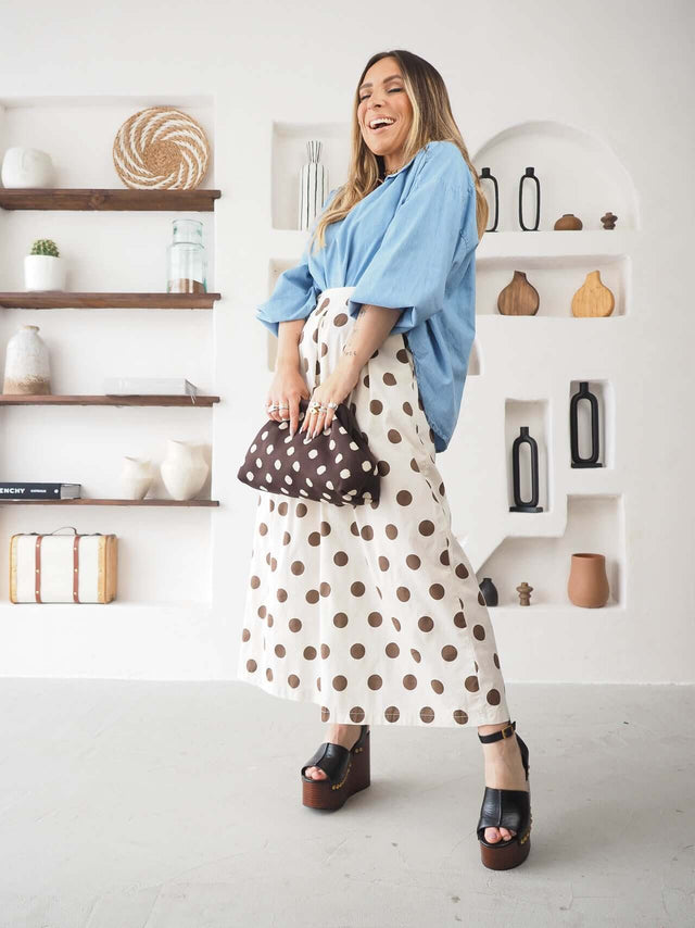 Mulher a sorrir com calças culote às bolas, blusa azul e sandálias altas, em ambiente moderno e descontraído.