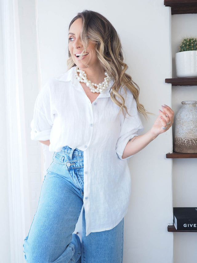 Mulher sorridente a usar camisa de linho branca, com colar de pérolas, jeans, em ambiente iluminado.