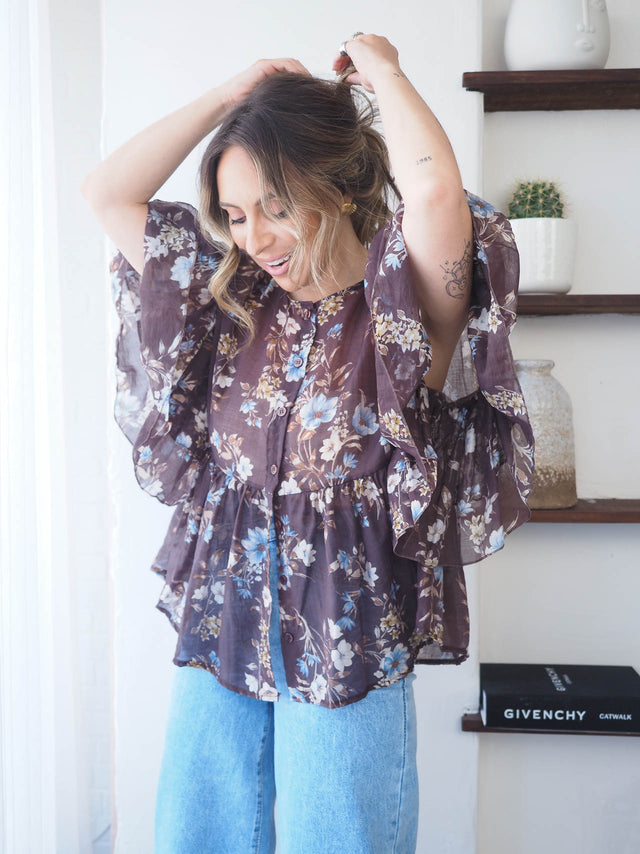 Modelo a sorrir com uma blusa florida semi-transparente, destacando o padrão floral e a leveza da peça.