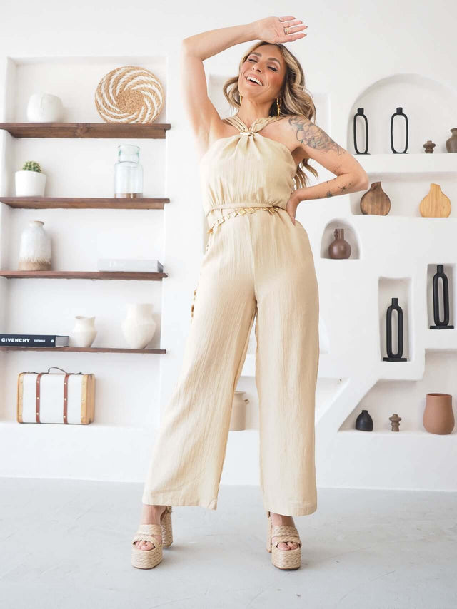 Macacão halter bege com cinto dourado, braço erguido, pose animada.