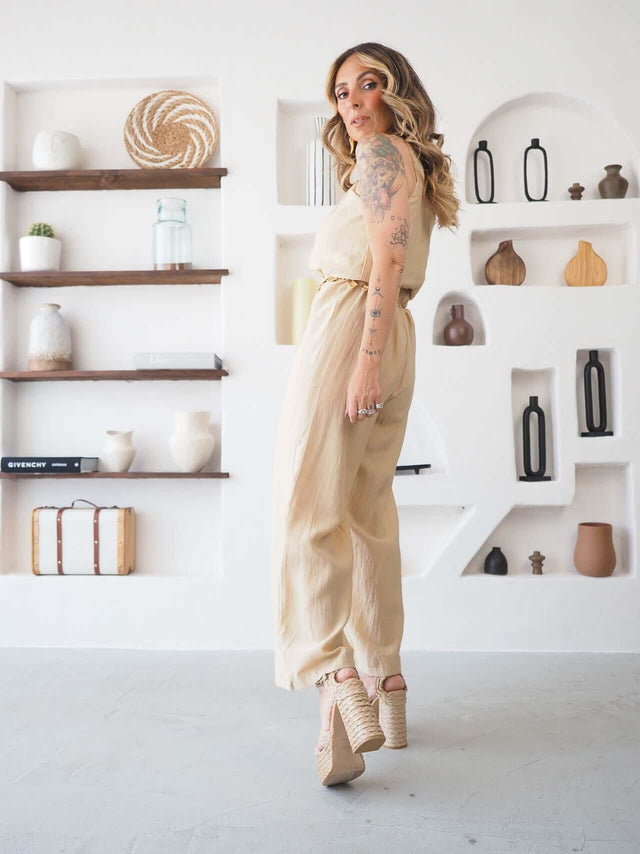 Macacão halter bege com cinto dourado, vista de costas com a modelo a olhar por cima do ombro.
