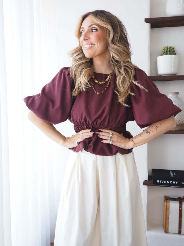 Blusa bordô de mangas balão, mãos na cintura, a sorrir.