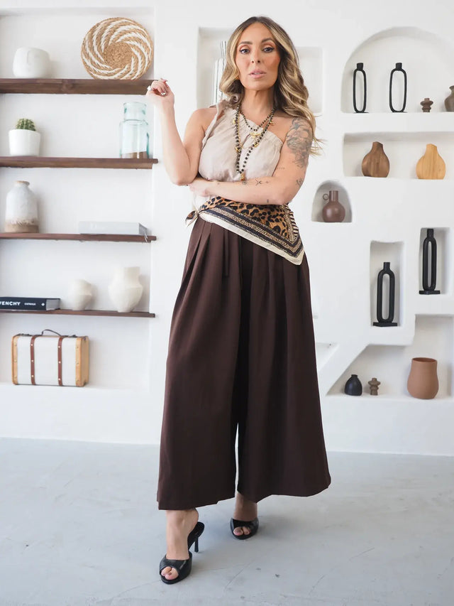 Mulher com calças pantalona culote castanhas e blusa clara, postura descontraída.